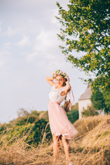  Beautiful model, slim blonde has a rest on a background of sunrise on the river near the tree and nature. White and pink dress, wreath of flowers. Air baths. Healthy lifestyle concept. 