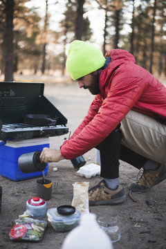 Man in warm clothing pouring water in cup while crouching on road