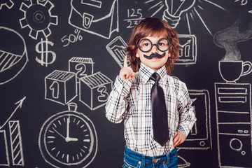 Little boy as businessman or teacher with mustache and glasses standing on dark background pattern. Wearing shirt, tie. Lifting a finger up