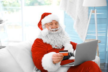 Santa Claus with credit card and laptop on couch at home