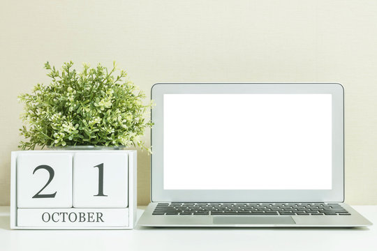 White wooden calendar with black 21 october word with white blank space at center of computer notebook on white wood desk and cream wallpaper textured background , selective focus at the calendar