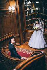 elegant gorgeous bride and stylish groom standing on wooden stairs in amazing old rich room. unusual wedding couple in retro style. luxury wedding concept. Man with long hair and piercing