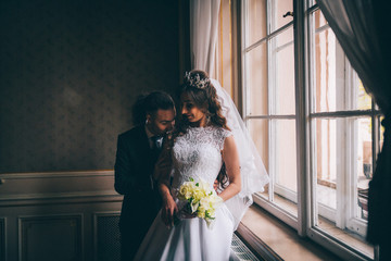 elegant gorgeous bride gently looking under veil at stylish groom standing at window light, holding calla bouquet. unusual wedding couple in retro style