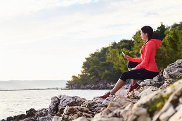 Fototapeta na wymiar Active young woman listening to music. Runner resting from routine exercise. Happy jogger relaxing at the beach after run, sitting on the stones and looking at the sea.
