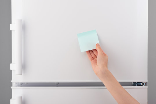 Female hand holding blank blue sticky paper note on refrigerator