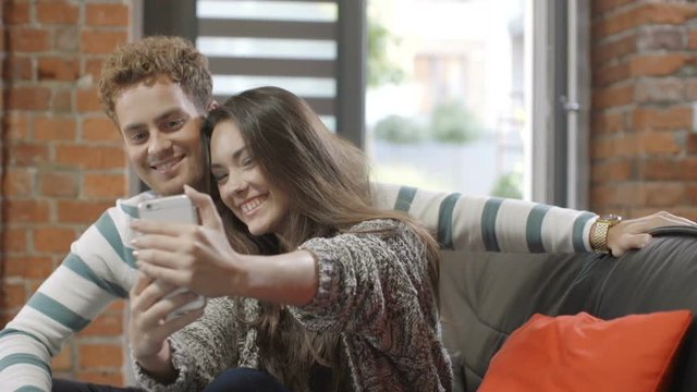 Attractive young couple making selfies with smart phone while sitting on a couch in a living room in modern loft interior. Happy young couple using smartphone at home.