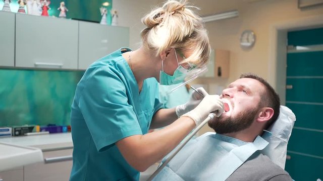 Female dentist examining teeth of male patient. Visit is in proffessional dental clinic. He is sitting on dental chair. He is young and has beard. Steadicam shot.
