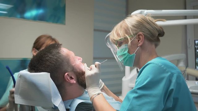 Female dentist drilling man's tooth with the help of an assistant. Visit is in proffessional dental clinic. He is sitting on dental chair. He is young and has beard. Steadicam shot.
