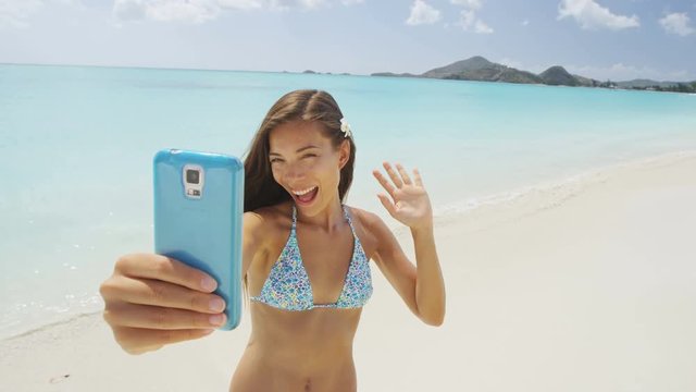 Phone girl using smartphone on beach waving hand saying hello hi on video chat having fun on beach vacation on summer travel holidays taking a selfie video. Sexy young bikini woman posing for camera.