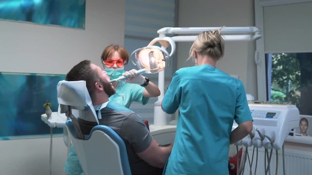 Dental assistant using dental curing lamp. Visit is in proffessional dental clinic. He is sitting on dental chair. He is young and has beard. Steadicam shot.
