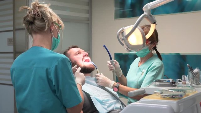 Dentist and assistant putting filling in the tooth. Visit is in proffessional dental clinic. He is sitting on dental chair. He is young and has beard. Steadicam shot.
