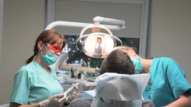Dentist puts filling in the tooth and assistant uses dental curing lamp. Visit is in proffessional dental clinic. He is sitting on dental chair. He is young and has beard. Steadicam shot.
