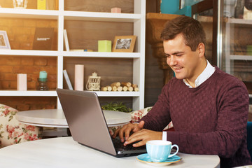 Happy young man smiling, as he works on his laptop to get all his business done early in the morning with his cup of coffee