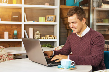 Happy young man smiling, as he works on his laptop to get all his business done early in the morning with his cup of coffee