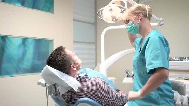 Dentist sits down and examining teeth of patient. Slider shot, right. Male patient on visti at young female dentist. Visit is in proffessional dental clinic. He is sitting on dental chair.
