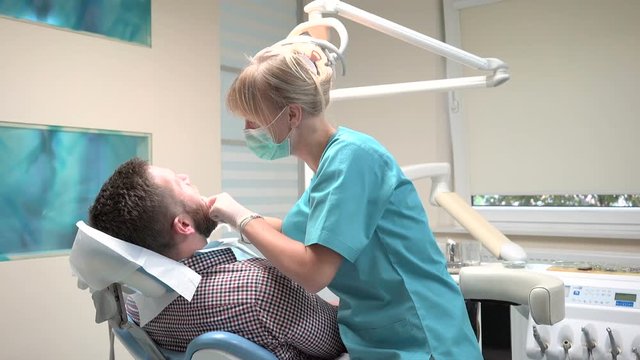 Female dentist checks up teeth of male patient. Male patient on visit at young female dentist. Visit is in proffessional dental clinic. He is sitting on dental chair. He is young and has beard.
