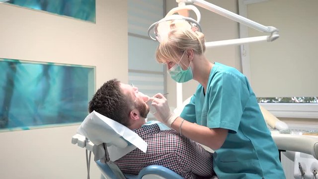 Male patient on visit at young female dentist. Visit is in proffessional dental clinic. He is sitting on dental chair. He is young and has beard.Dentist examining patient's teeth and talking.