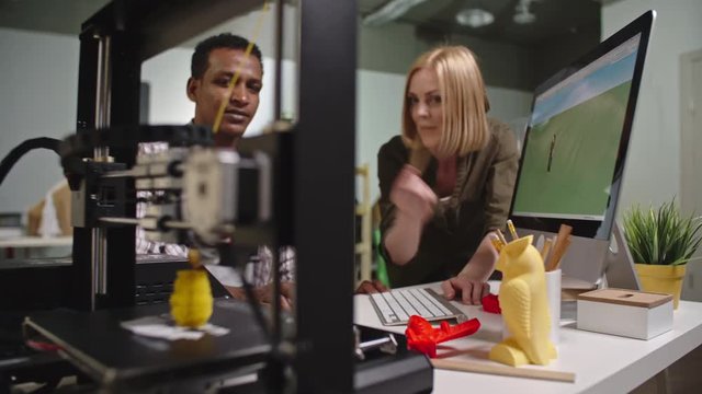 Rack focus from female worker looking at computer monitor and discussing 3d model with African colleague to working FDM 3d printing machine 
