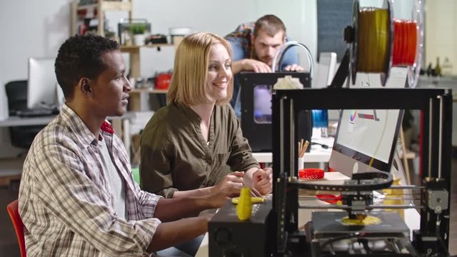 Side view of African engineer sitting behind computer with female executive and showing her 3d printed detail, colleague working in background 