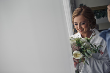 Stunning blonde bride with delicate hair smiles holding a weddin