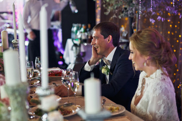 Groom smiles holding his head with a hand and sitting behind a b