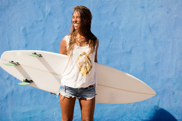 happy surfer girl with surfboard in front of blue wall