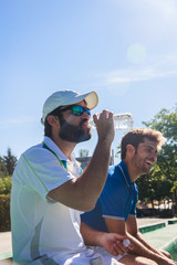 Two professional players hydrating after a hard game of tennis. They are friends and are sitting resting for the game. They are happy and laughing. 