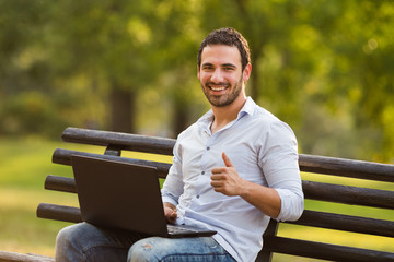 Fototapeta premium Young businessman is sitting at the park,using laptop and showing thumb up.