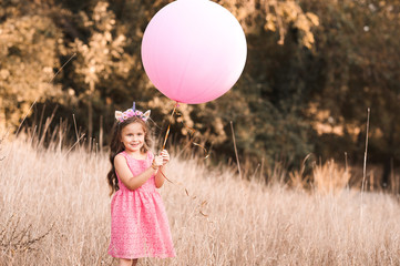 Stylish baby girl 4-5 year old holding big balloon wearing trendy pink dress in meadow. Playful. Birthday party.