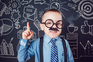 Little boy as businessman or teacher with mustache and glasses standing on dark background pattern. Wearing shirt, tie. Lifting a finger up