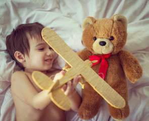 Happy kid playing with wooden toy airplane and Teddy bear in bed