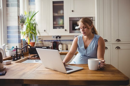 Pregnant woman using laptop while having coffee in kitchen