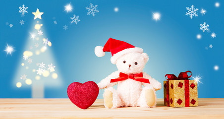 Christmas card for kids. Stuffed teddy bear with Santa Claus hat, present, and heart on the wood table. Christmas tree in the background. 