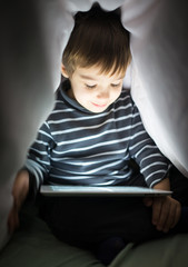 Cute little boy alone with tablet computer under blanket at nigh