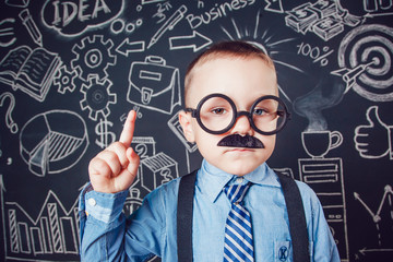 Little boy as businessman or teacher with mustache and glasses standing on dark background pattern. Wearing shirt, tie. Lifting a finger up