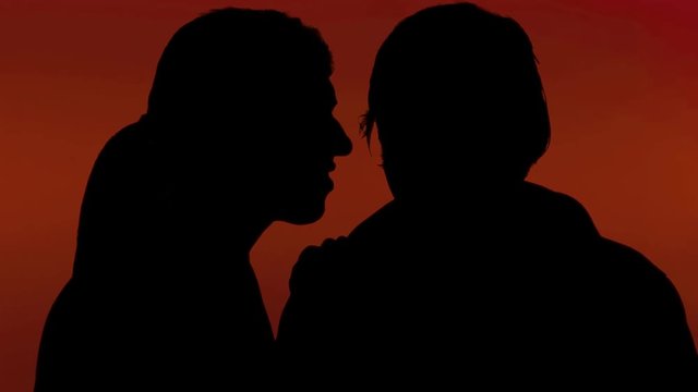 A woman whispering a secret in the ear of a man. Silhouette shot, red background.
