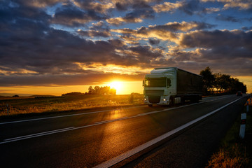 White truck driving on asphalt road along the corn field during a dramatic sunset.