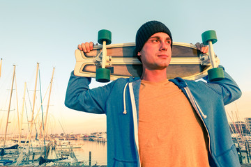 Thoughtful man holding skateboard on shoulders looking at sunset with boat on background