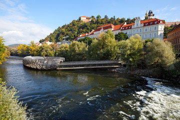 Fototapeta na wymiar River Mur with the artificial floating platform Mur island (Murinsel) in the middle and old buildings on the river bank. Graz, Austria.