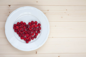 Heart out of fresh raspberries on a white plate. Light wood table. 
