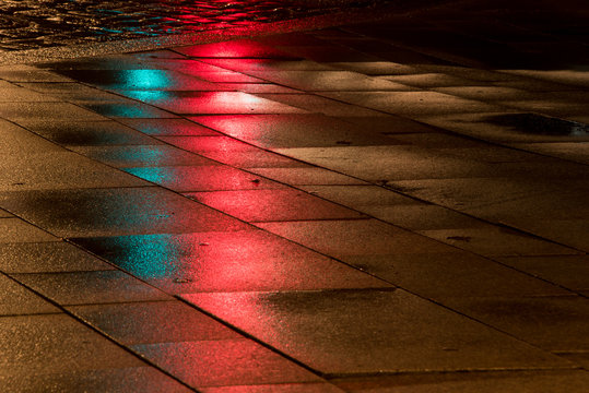 leuchtende Steinplatten im Regen bei Nacht mit Spiegelung von Ampellicht und Stra&szlig;enlaternen