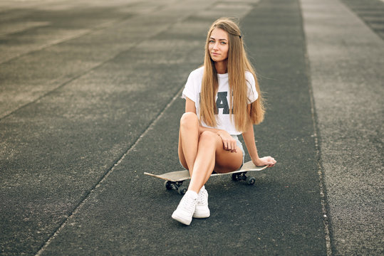 Beautiful young and sexy girl in a white T-shirt and short shorts sitting on a skateboard