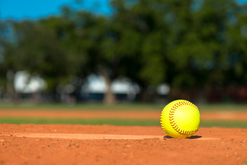 Bright fluorescent neon day-glo yellow softball on pitchers mound with red dirt field grass and...