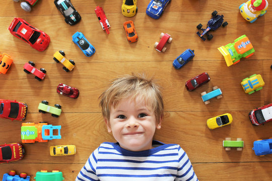 A young boy on a wood floor with all his cars and toys around him. Picture taken from above him, with a focus on his face and blurry toys