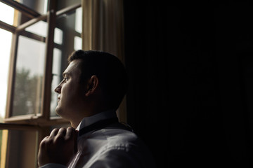 Brunette groom is standing next to the window