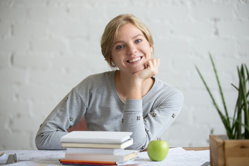 Portrait of a young smiling student girl sitting at the desk with books and green apple, propping...