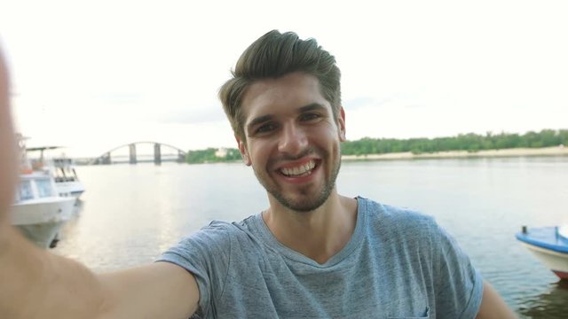 Cheerful selfie. Cheerful young man in shirt holding mobile phone and making photo of himself while standing in city river