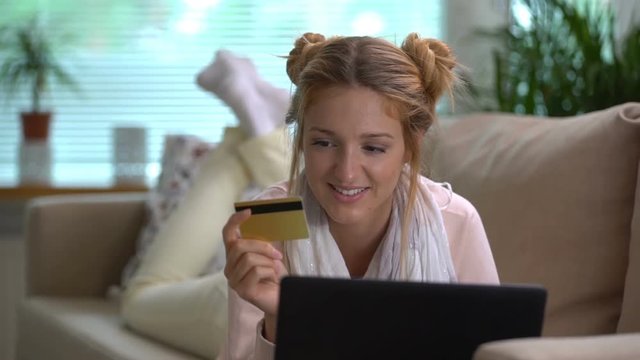 A young woman uses a golden credit card online lying on the couch. Slow motion.