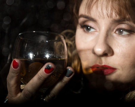 mysterious young woman with a glass of wine posing behind transparent glass covered by water drops. on a dark background
