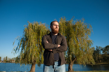 serious young man with a beard stands on a background of trees and lake